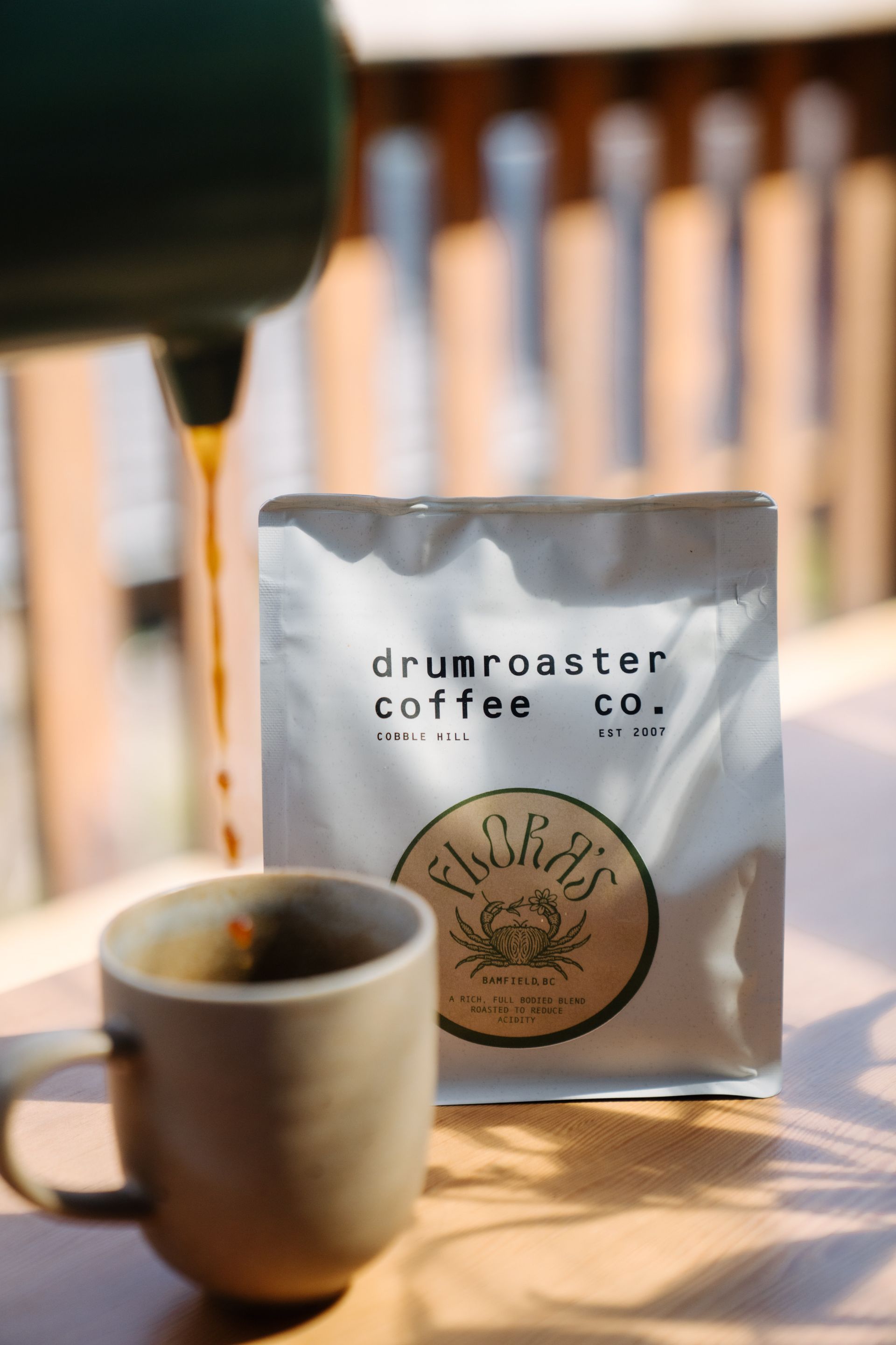 Coffee being poured from a green vessel into a ceramic mug next to a white Drumroaster Coffee Co. bag on a wooden table.