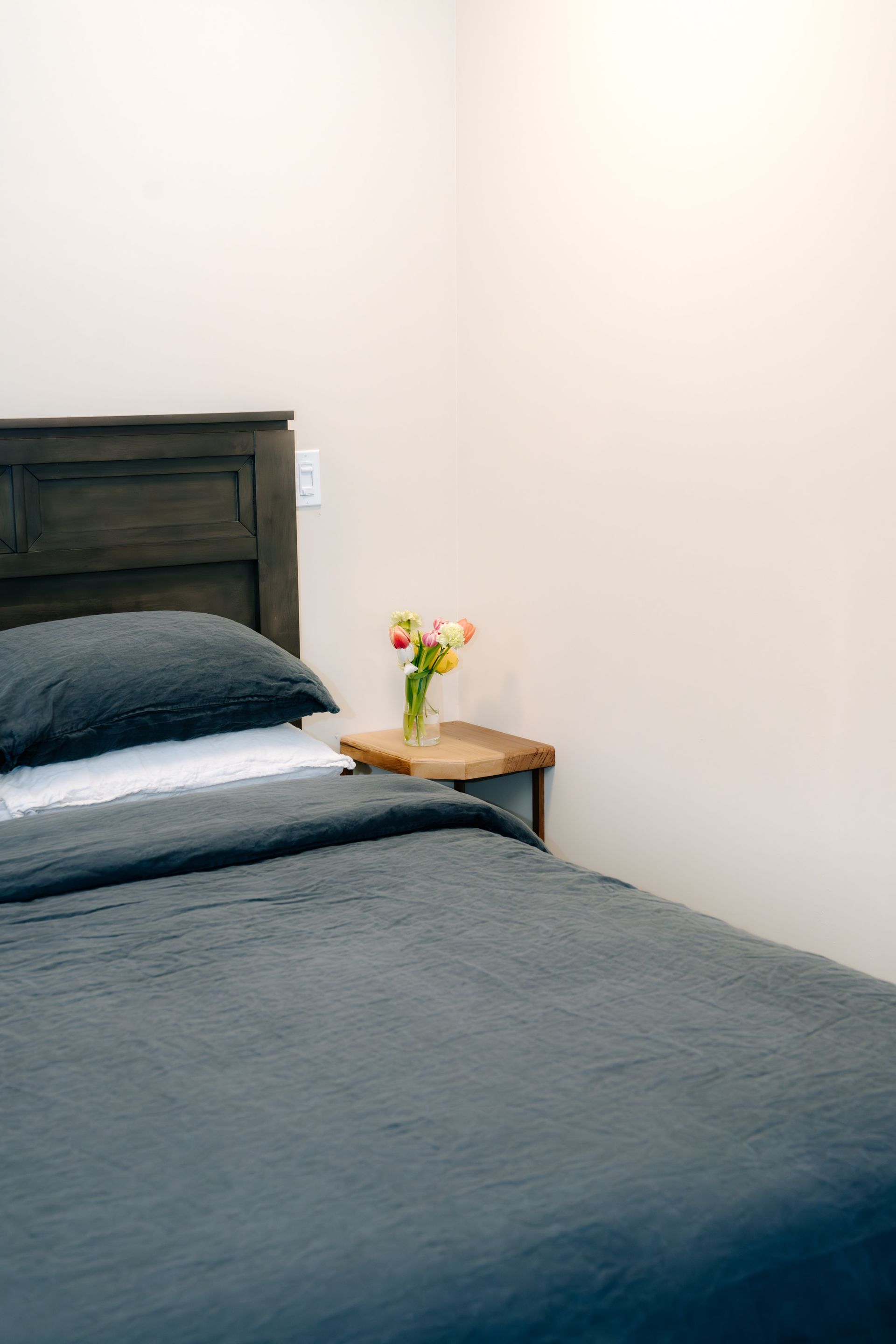 A bed with dark gray bedding and a wooden headboard, next to a small wooden side table holding a bouquet of flowers.