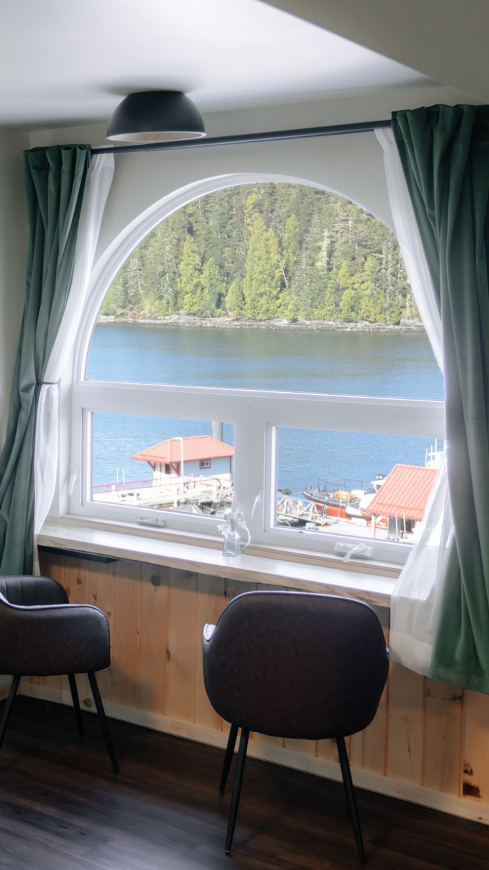 Two dark chairs sit before an arched window overlooking a blue bay with forested hills and small buildings.