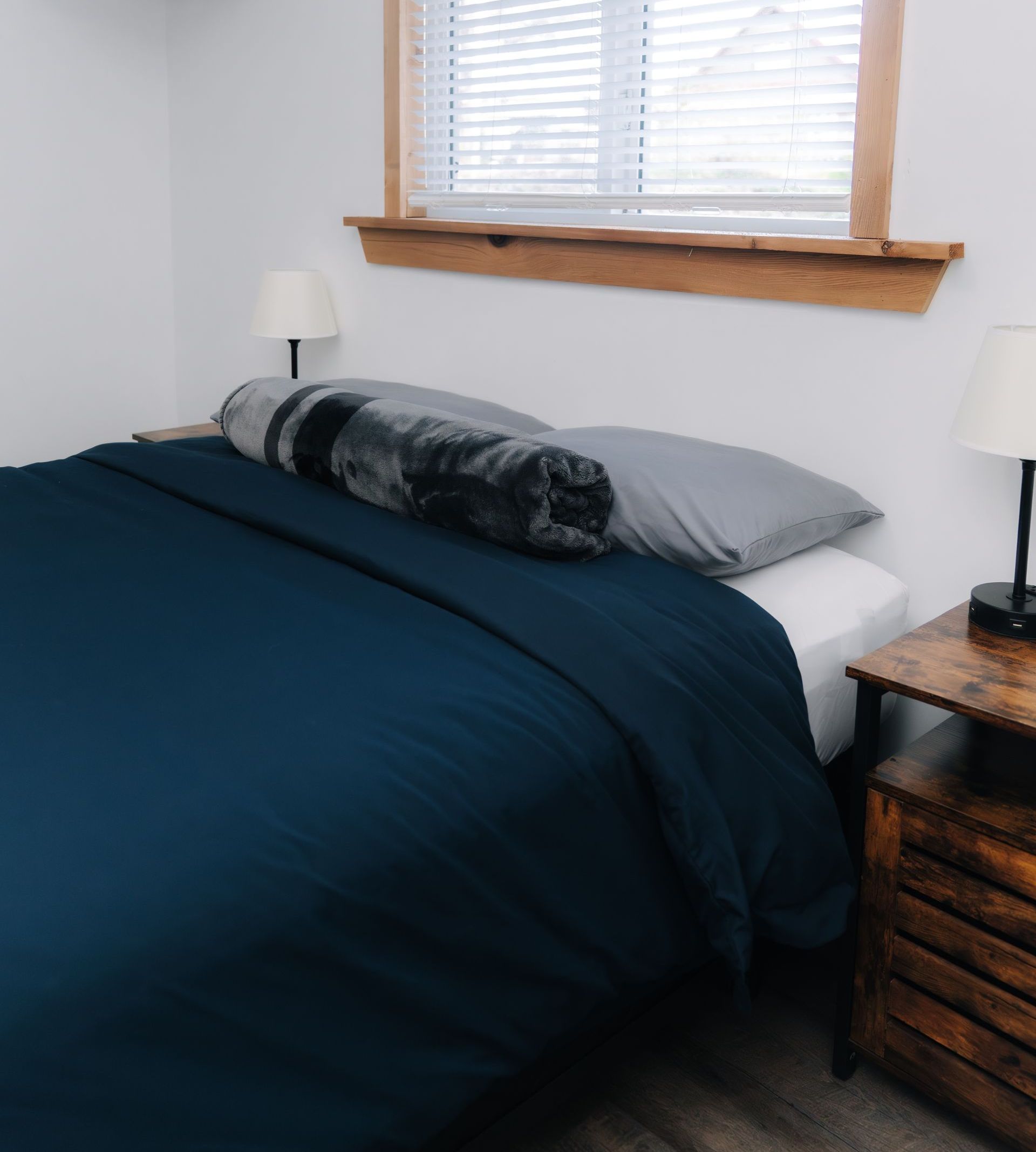 A bed with a dark blue comforter and gray pillows sits beneath a window with wooden trim and two bedside lamps.