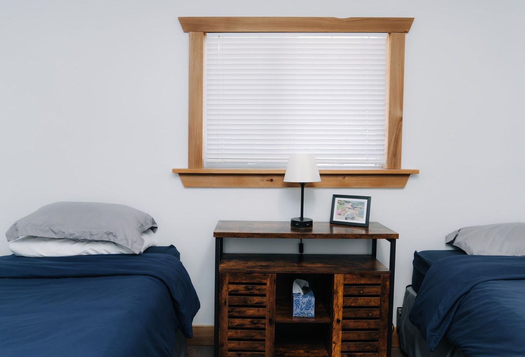 Two beds with blue bedding and grey pillows, separated by a wood cabinet with a small lamp and picture frame.