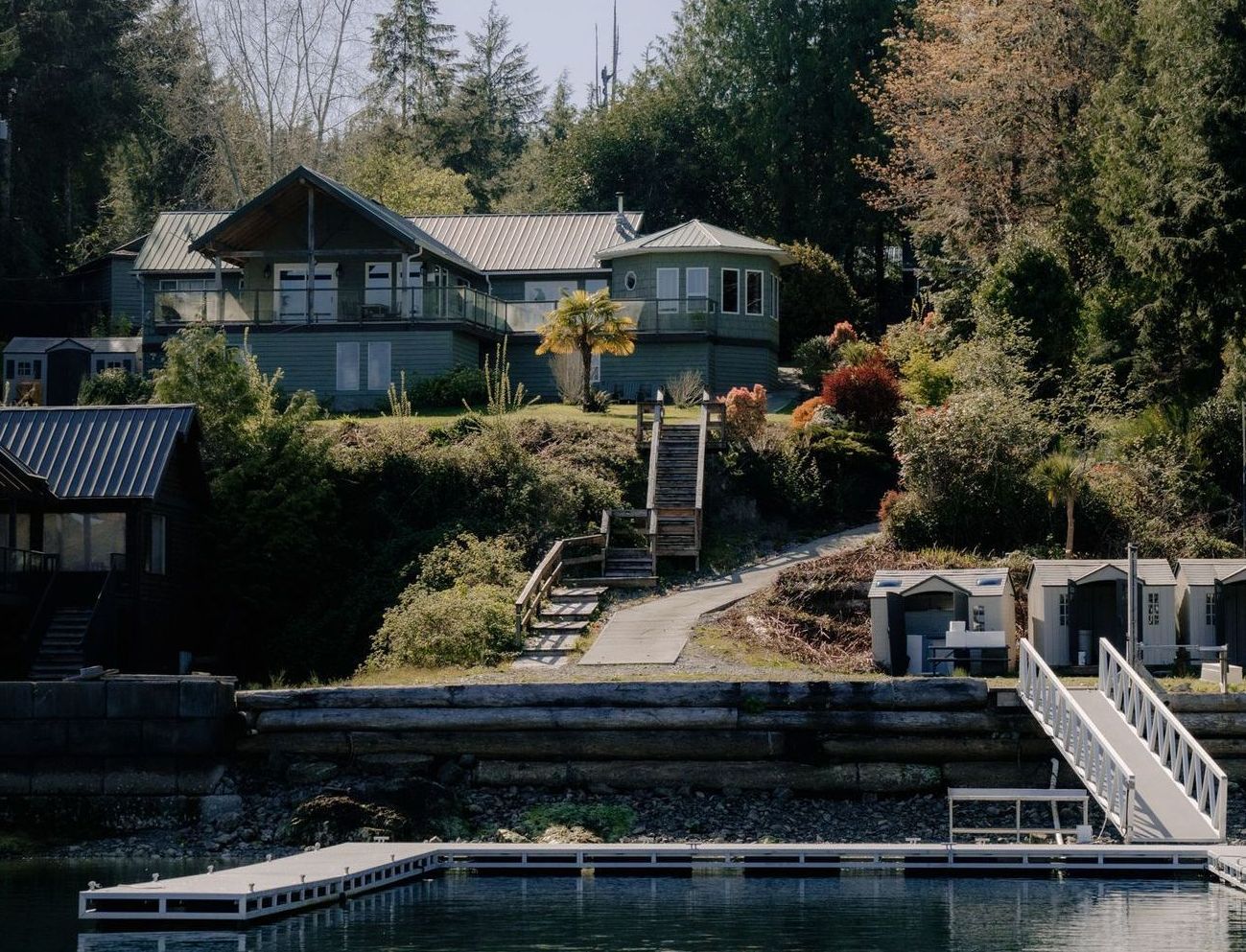 A metal ramp leads down to floating docks at a coastal harbor with wooden structures and lush, forested hills behind.