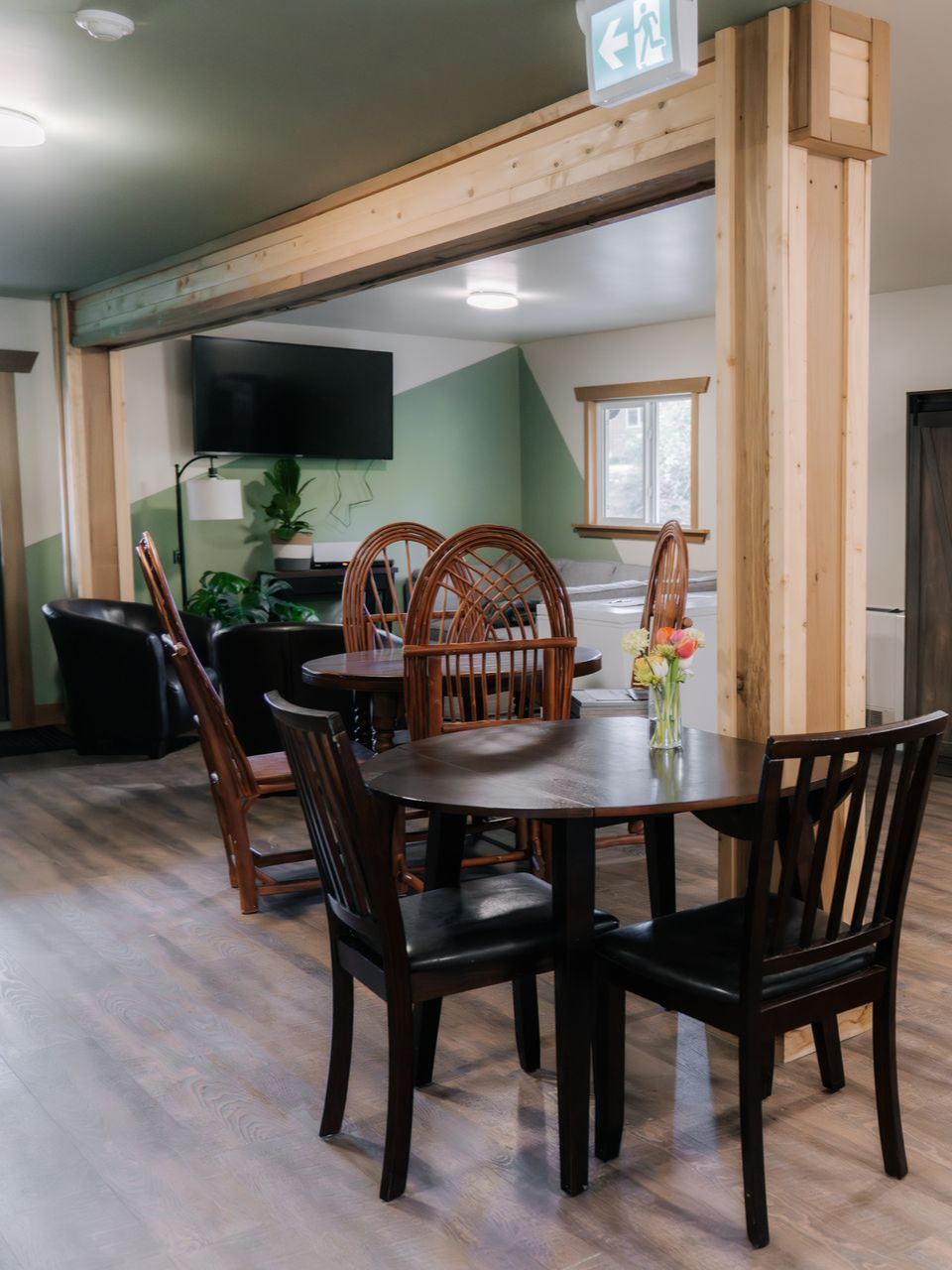 A dining area with a wooden table, dark chairs, and a structural beam, leading to a lounge with a TV and green wall paint.