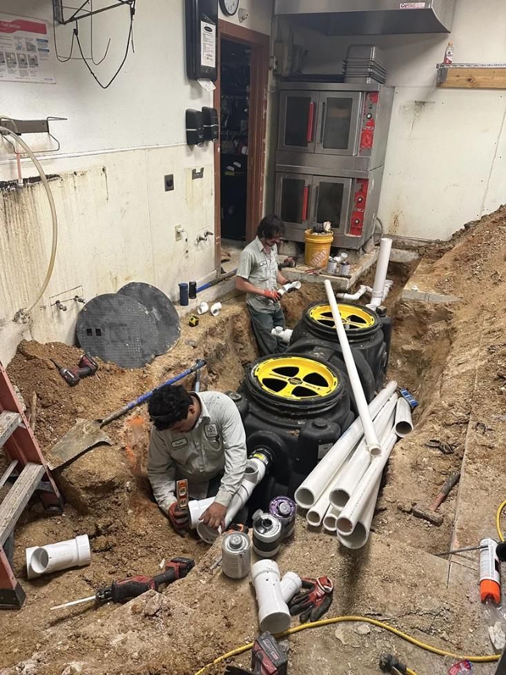 Worker kneeling in a trench amid pipes and cables at an indoor construction site