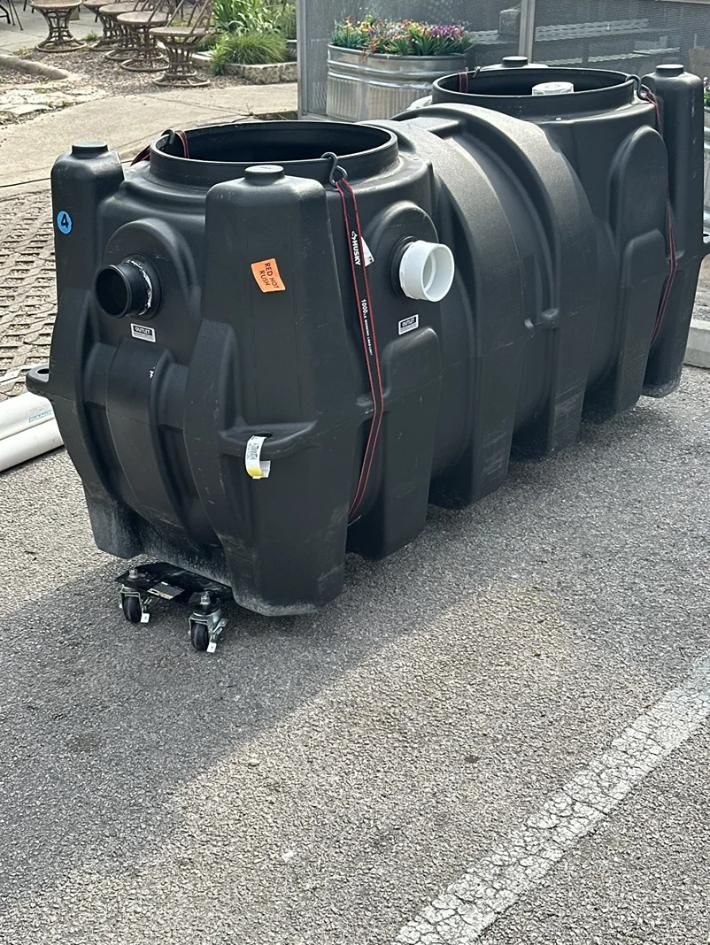 Large black plastic water tank on pavement, with pipes and fittings attached.