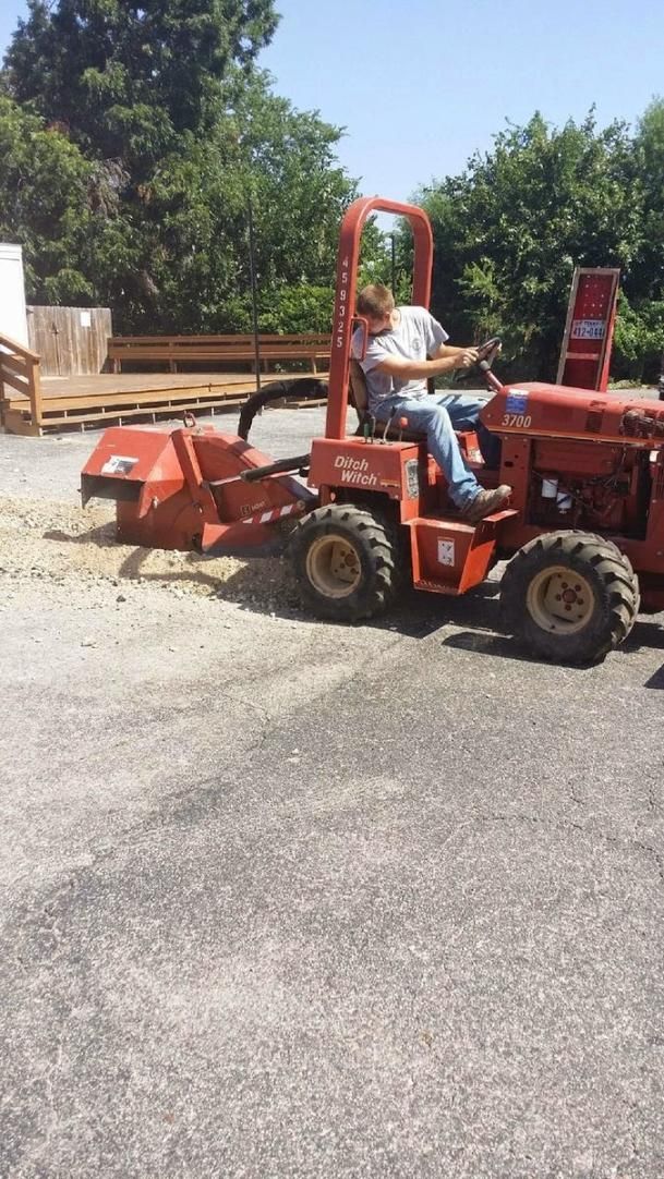 Red forklift carrying a worker in a gravel lot near stacked lumber and trees.