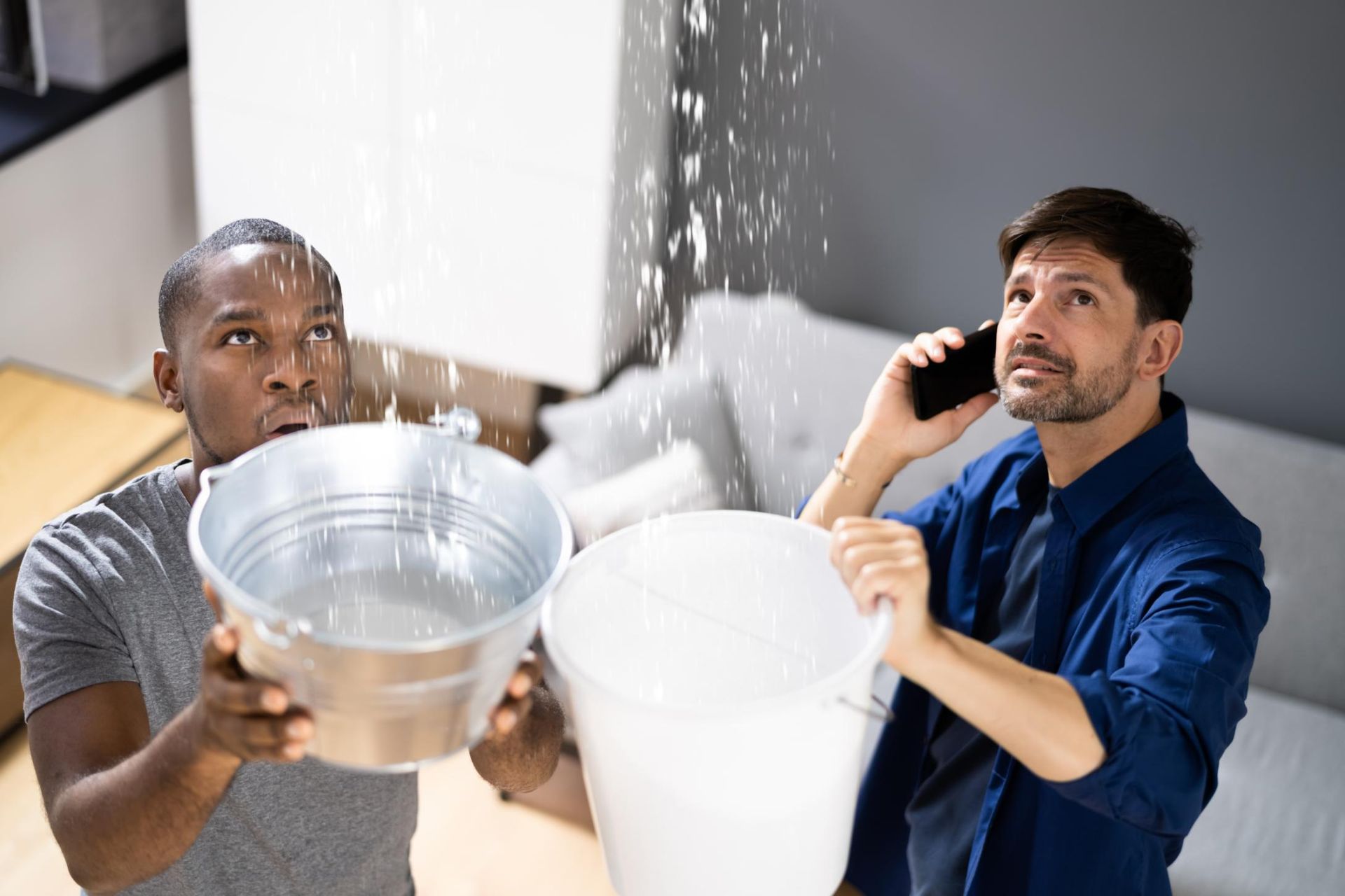 Two men catch water from a leaking ceiling with bowls, one on a phone, in a home interior