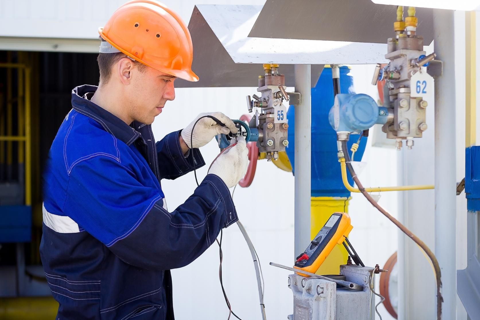 Worker in orange hard hat inspecting industrial equipment with a handheld device in a factory setting