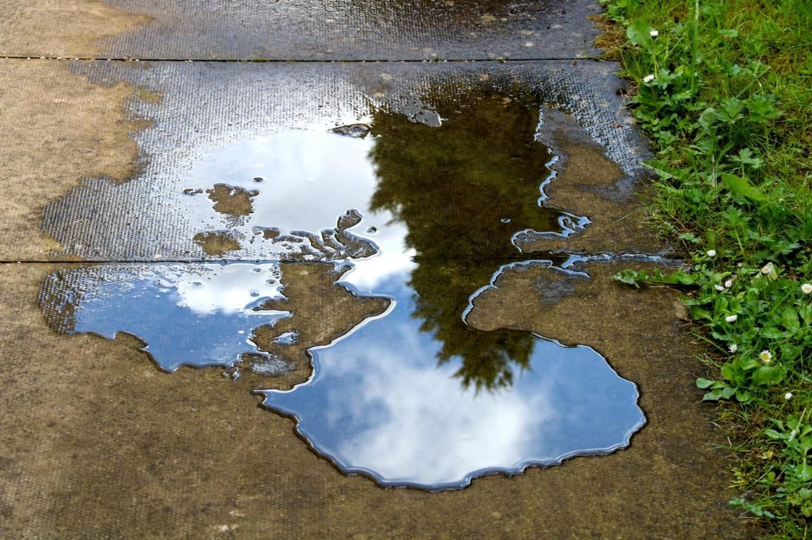 Puddles reflecting cloudy sky on a wet paved path beside grass and greenery