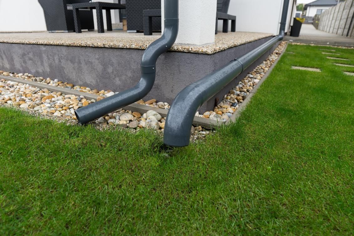 Black downspout pipes draining from a house onto gravel edging beside a green lawn.