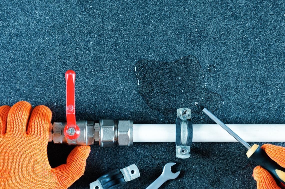 Orange-gloved hands working on a white pipe with fittings and a red valve on a dark blue floor