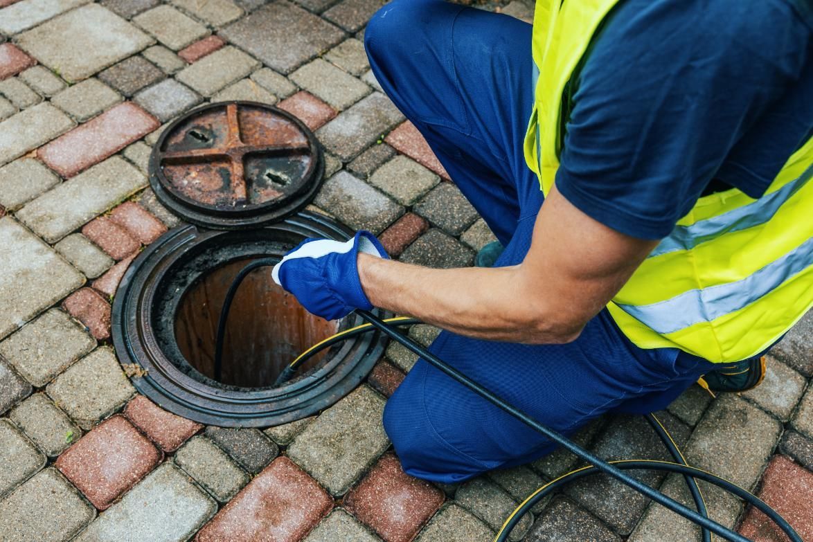 Worker in blue gloves and neon vest inspecting an open manhole on a paved street