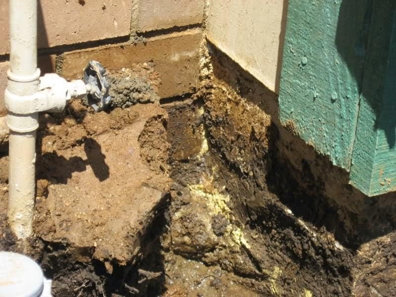 Cracked soil and mud at the base of a building near a white pipe and green siding
