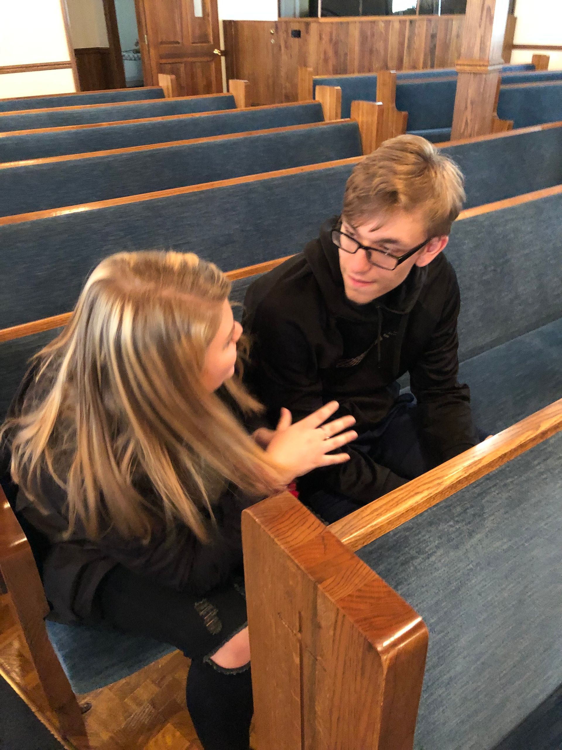 Two people talking in a wooden church pew, one gently touching the other's arm.