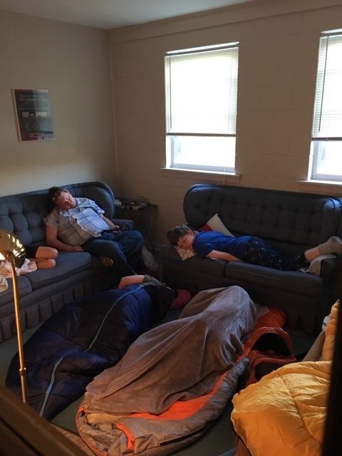 Two people resting on couches in a living room with blankets and two bright windows.