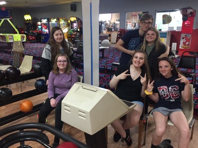Five friends pose together at a bowling alley, smiling near a lane and arcade games.