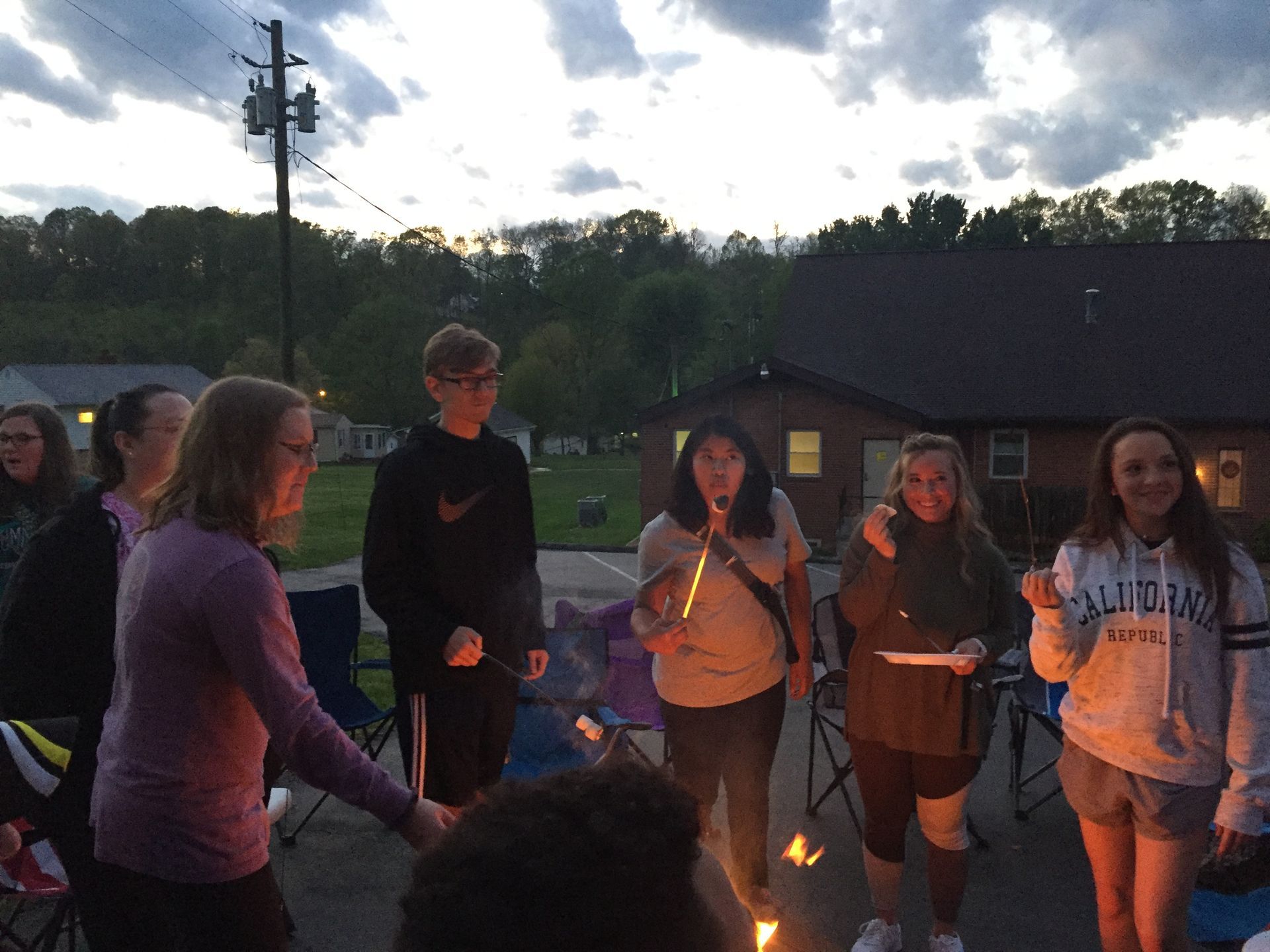Group of people holding candles outdoors at dusk near a campfire, standing by a building and utility pole.