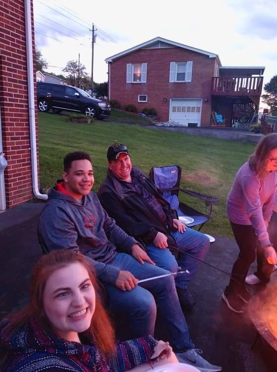 People sitting around a fire pit in a backyard at dusk, smiling and chatting with a house in the background