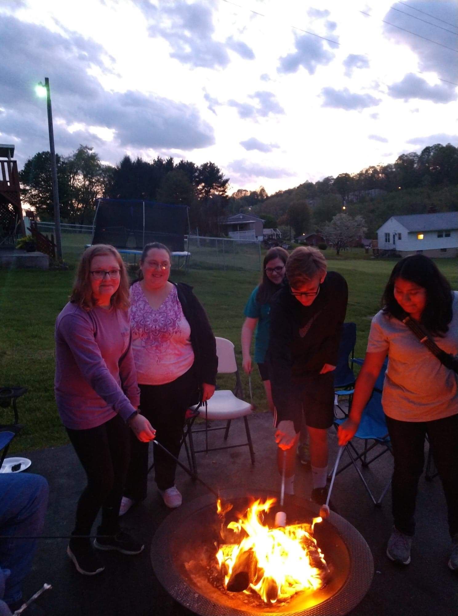Five people stand around a campfire at dusk in a grassy field, with houses and trees in the background.