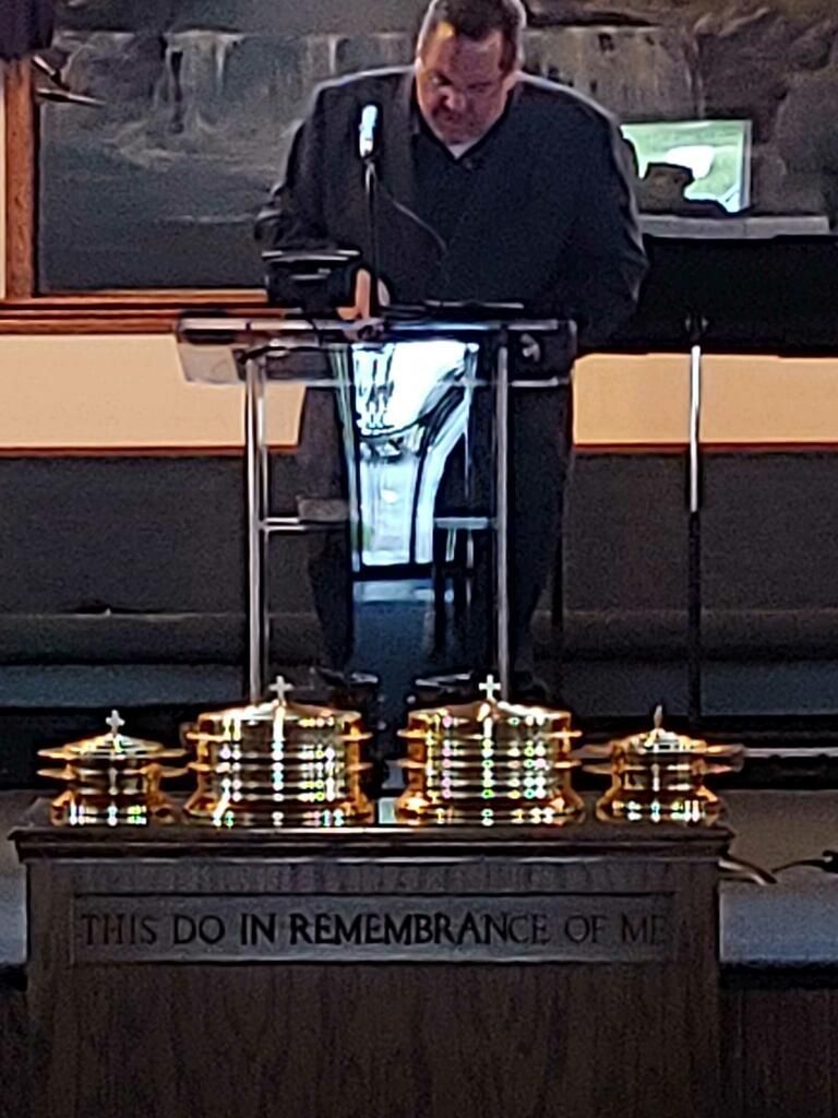 Person speaking at a podium in a church, with communion trays and “This do in remembrance of me” on the front.