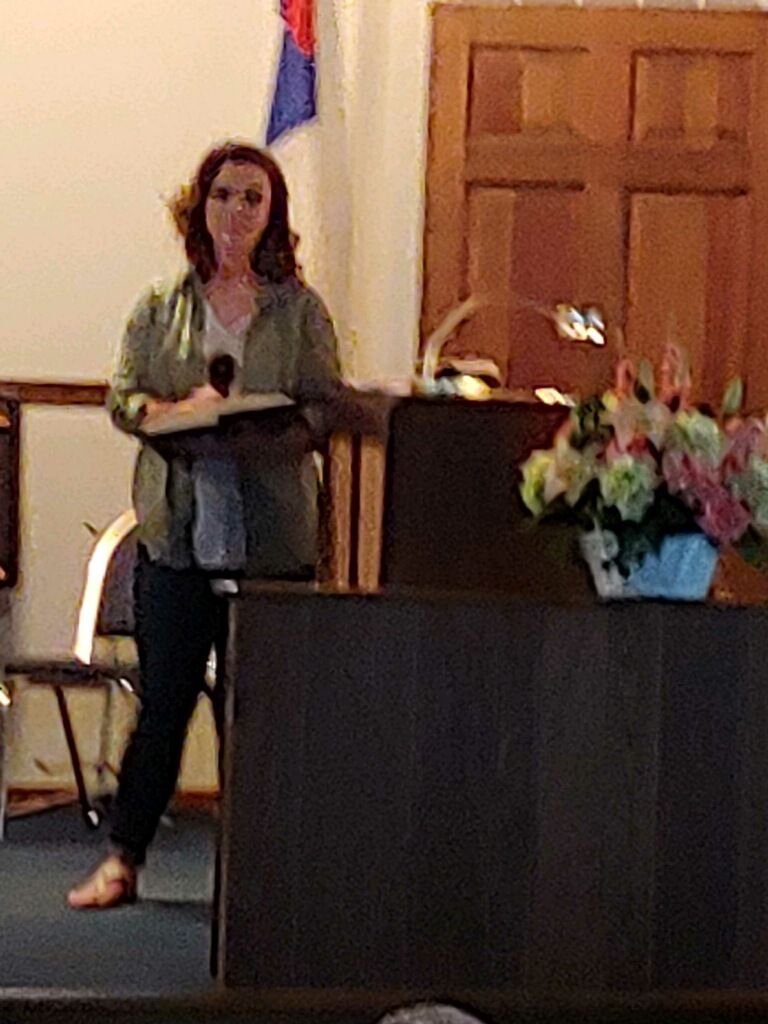 Woman standing at a podium holding a microphone, with flowers on the lectern in a room with wooden doors.