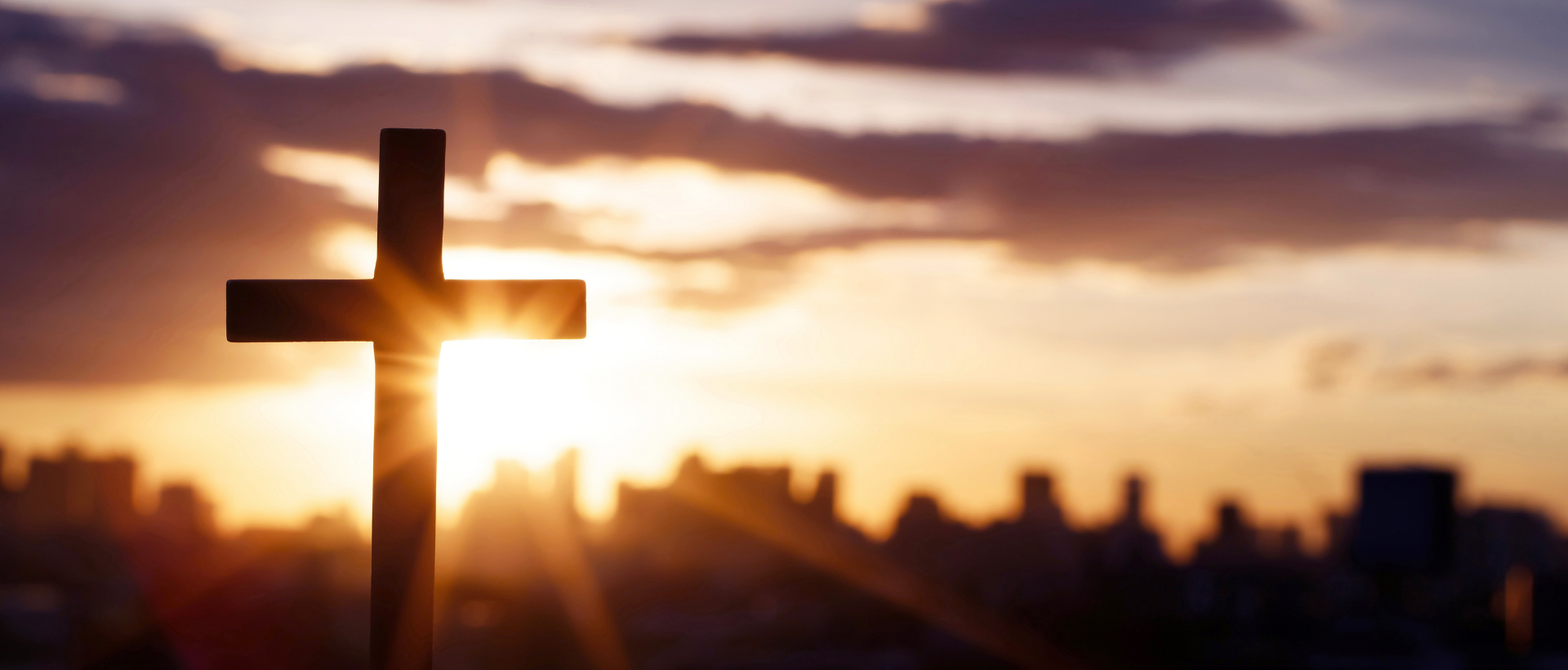 View Our Beliefs Silhouetted cross at sunrise with warm orange sky over a blurred city skyline