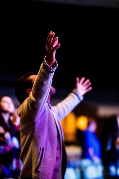 Learn More About Our Beliefs Person in a gray sweater raising both hands in a dimly lit crowd at an event