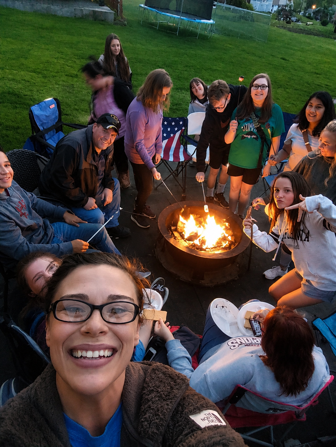Group of friends gathered around a backyard fire pit at dusk, smiling and taking a selfie