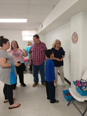 People chatting in a bright hallway near a blue-covered table with flowers and a wall emblem.