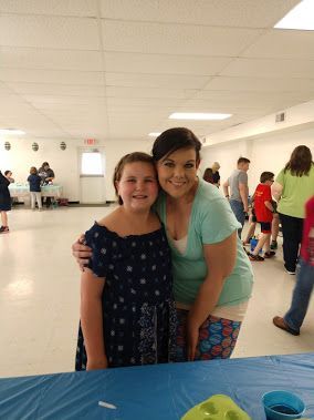 Two people smiling and hugging in a bright community hall with tables and people in the background