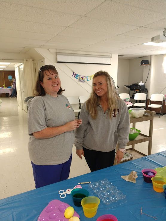 Two women smiling beside a craft table with cups, scissors, and supplies in a bright room.