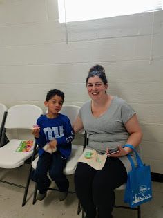 Two people sit on chairs in a bright room, smiling and holding small items; one has a blue tote bag.