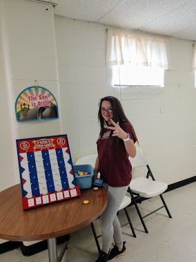 Person posing in a room beside a table with a patriotic board game and a window curtain backdrop
