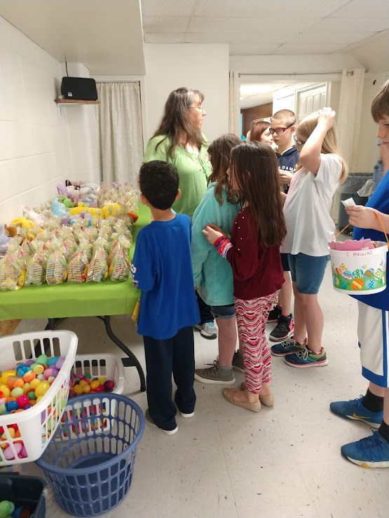 Children gather in a hallway by tables of colorful Easter eggs and baskets.