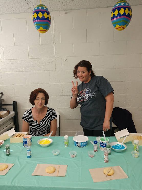 Two people at a table with plates and cups, one smiling and flashing a peace sign under colorful hanging balloons.