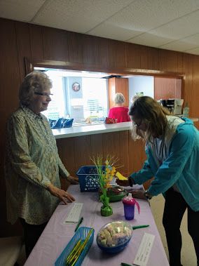 Two women arranging items on a table in a small community room with a service counter.