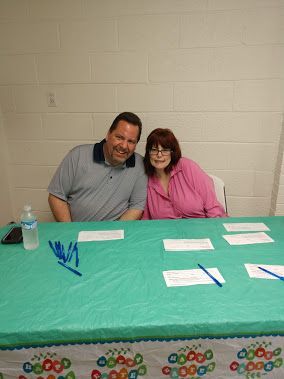 Two people seated behind a teal-covered table with papers and pens at a community event