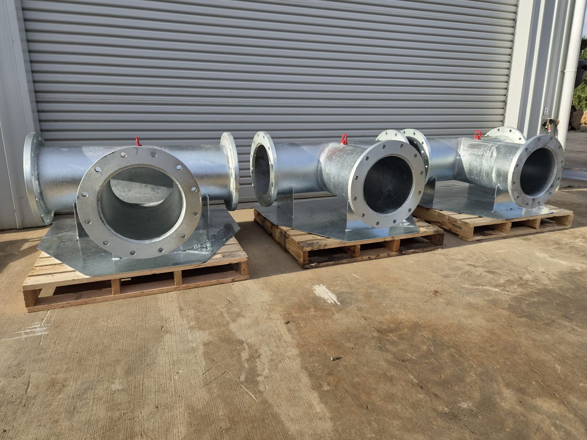 Three large silver industrial metal duct housings on wooden pallets outside a gray roll-up door — Forcequip Hire In Glanmire, QLD