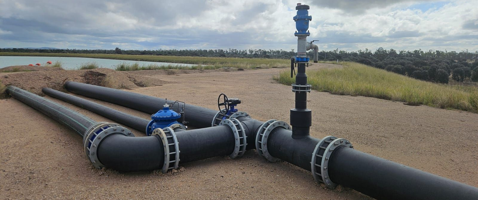 Large Black Industrial Pipes With Blue Fittings Running Across a Gravel Field Under a Cloudy Sky - Forcequip Glanmire QLD