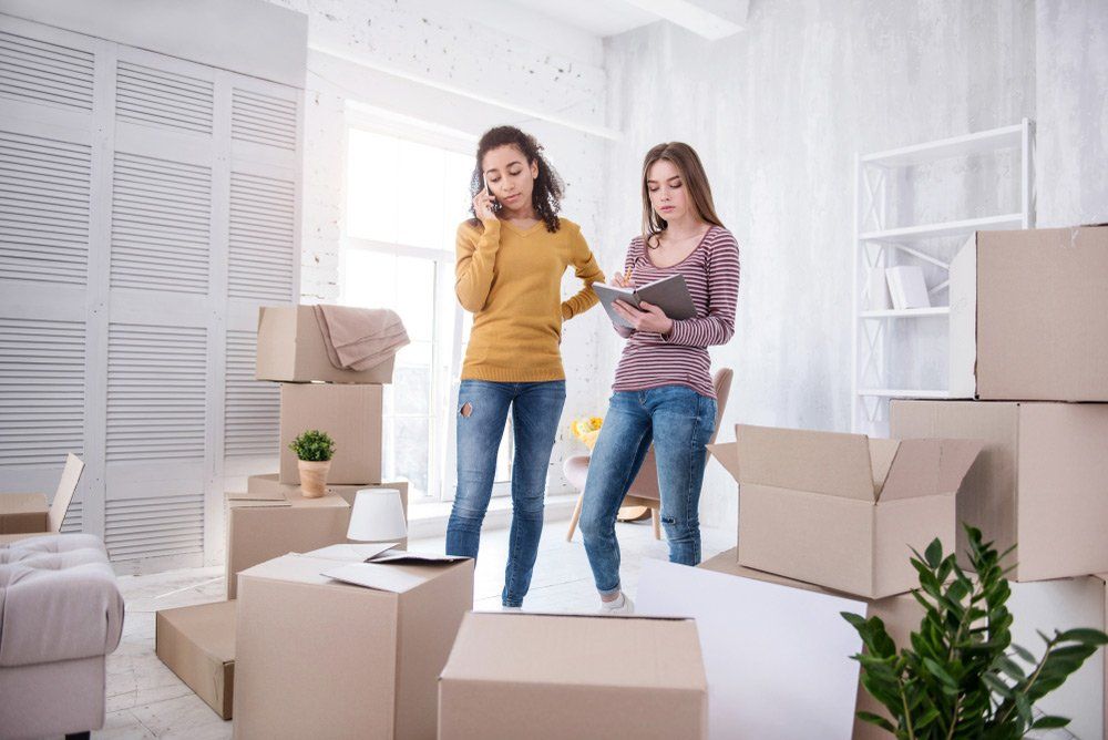 Two women standing in a room surrounded by moving boxes, reviewing a checklist.