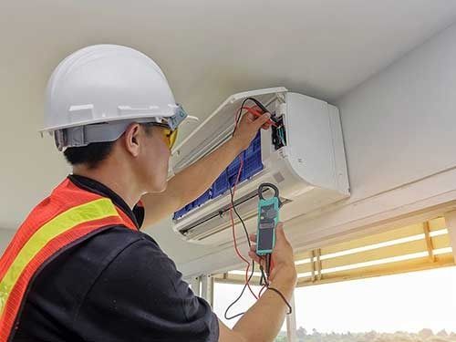 Un trabajador con casco inspecciona un aparato de aire acondicionado con un multímetro en una unidad montada en la pared.
