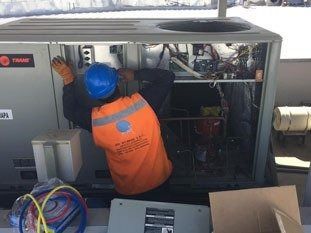 Técnico con chaleco naranja y casco azul inspeccionando un equipo de climatización con el panel abierto.