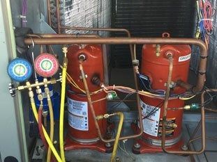 Dos compresores de climatización rojos con manómetros y tuberías de cobre en una sala de máquinas.