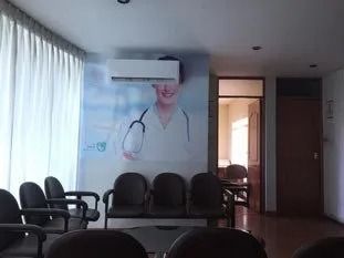 Sala de espera con sillas negras, grandes cortinas en las ventanas y un mural de un médico con un aire acondicionado encima.