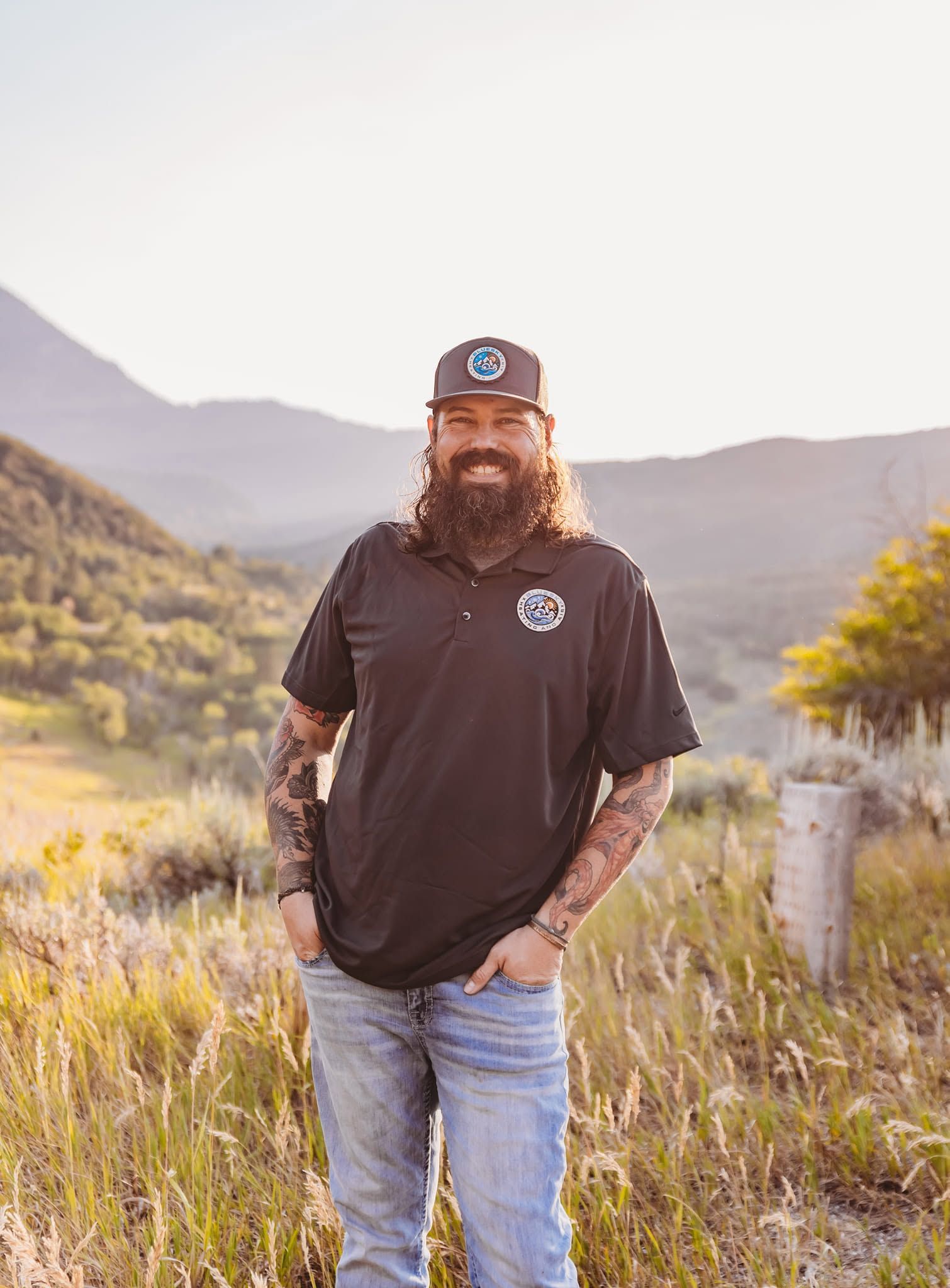 Bearded man in black shirt and cap standing in a grassy mountain valley