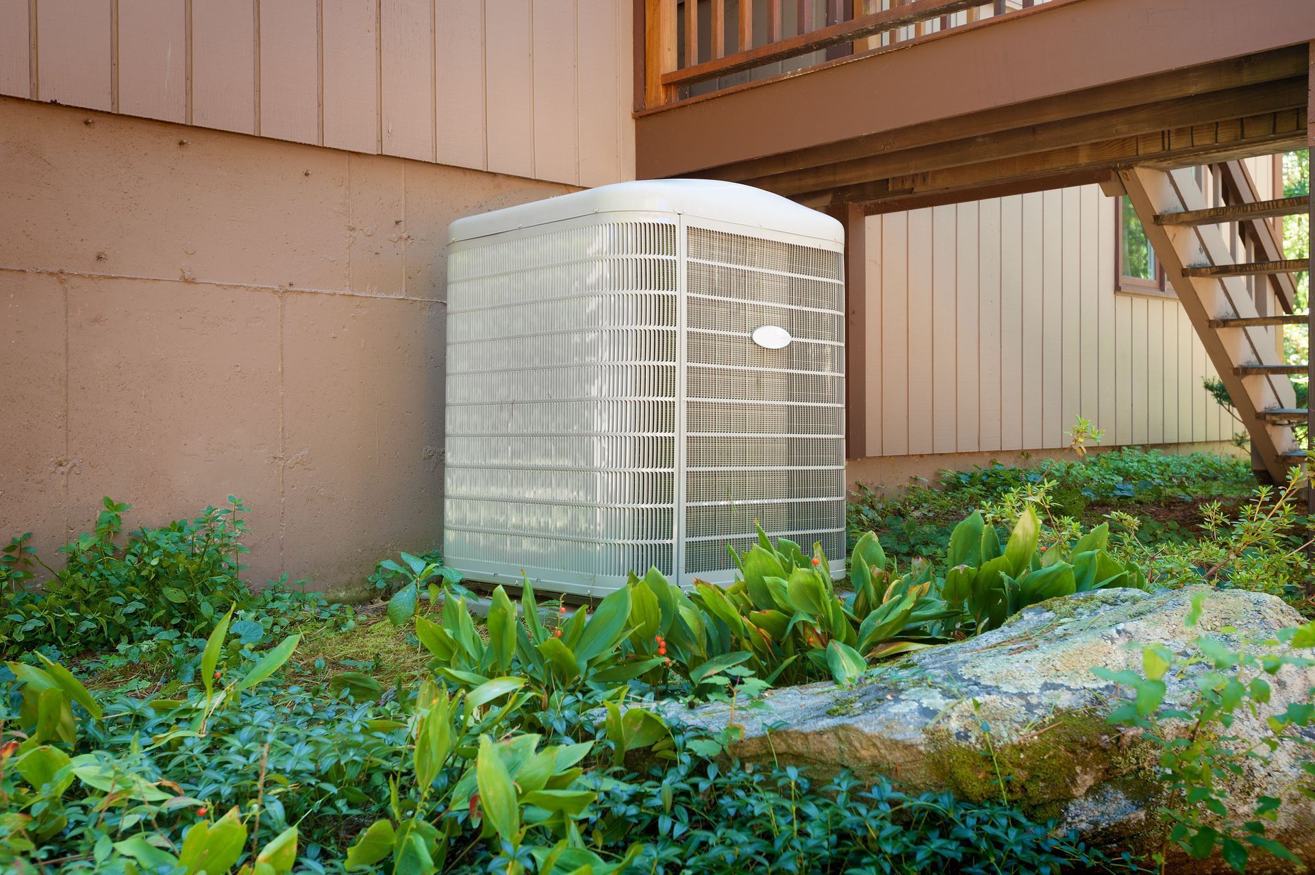 White air-conditioning unit beside a building, surrounded by green plants and a wooden deck stairs