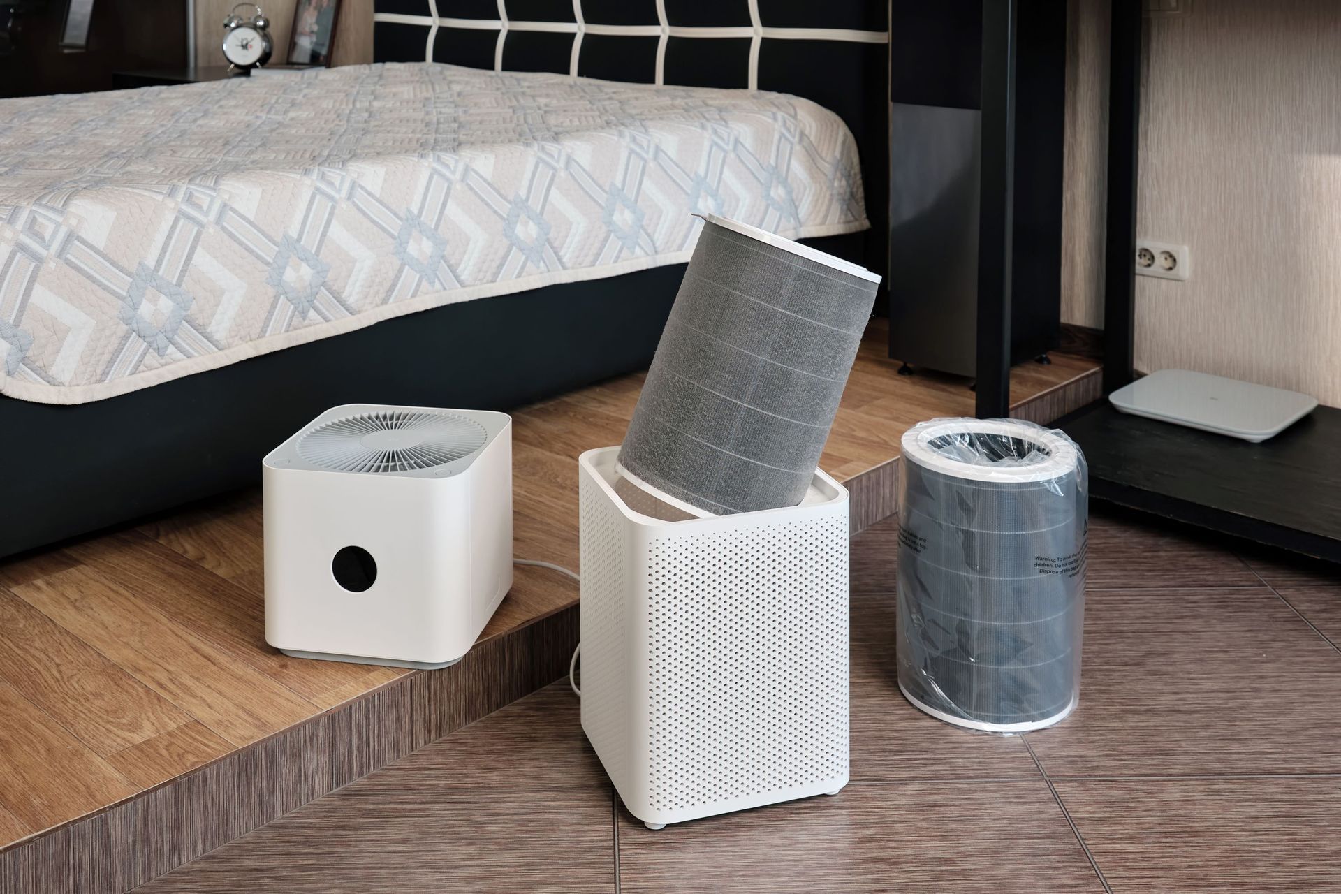 Three small air purifiers and filters on a wooden floor beside a bed