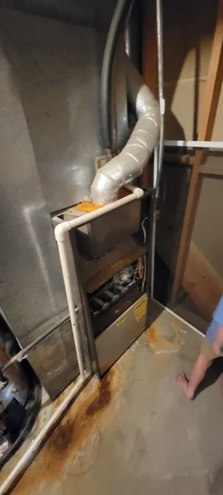 Open furnace with metal ducting in a basement utility area, with a person’s leg visible at the right edge