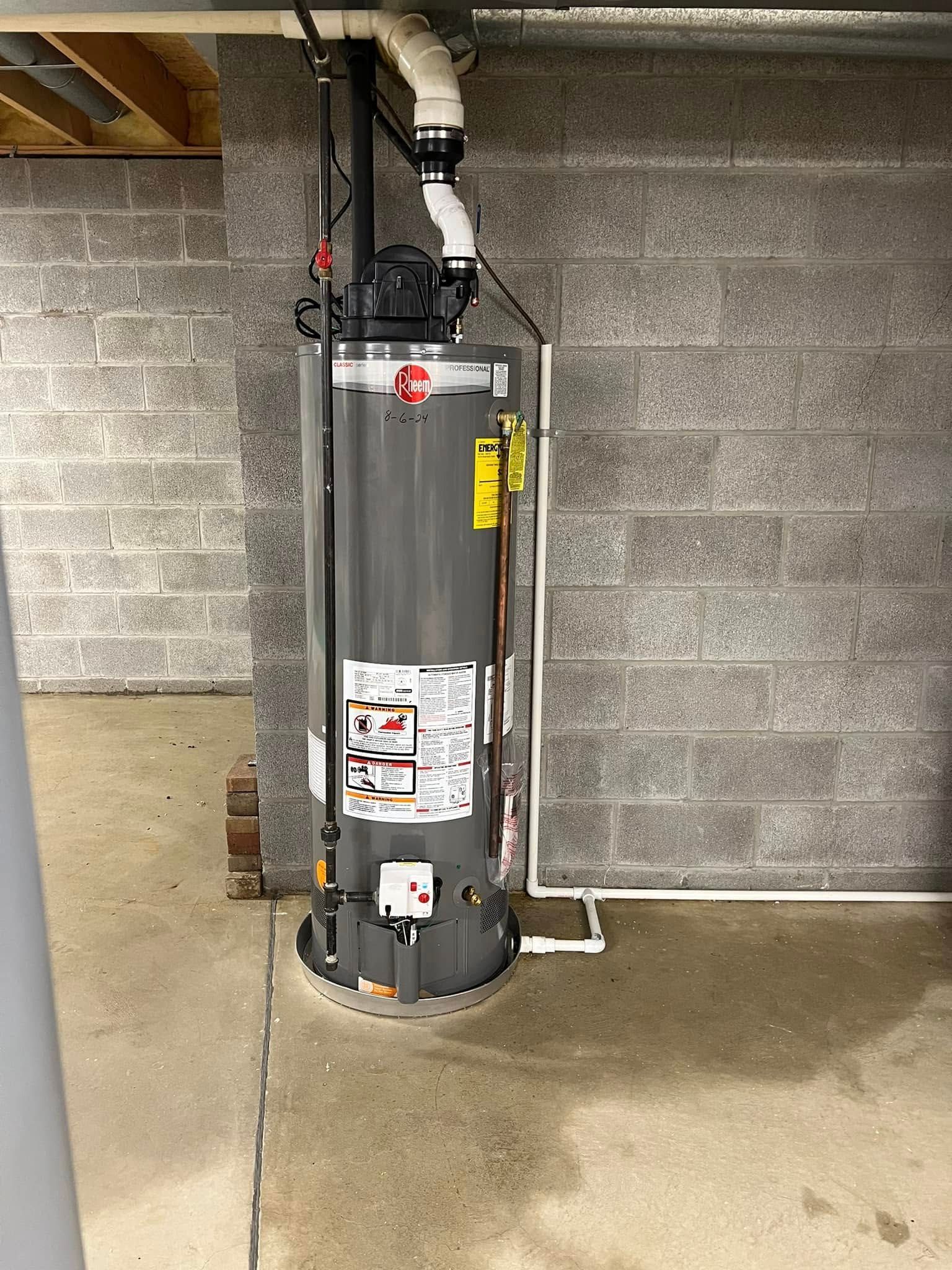A gray Rheem water heater standing on a concrete basement floor against a cinder block wall, with white piping above.