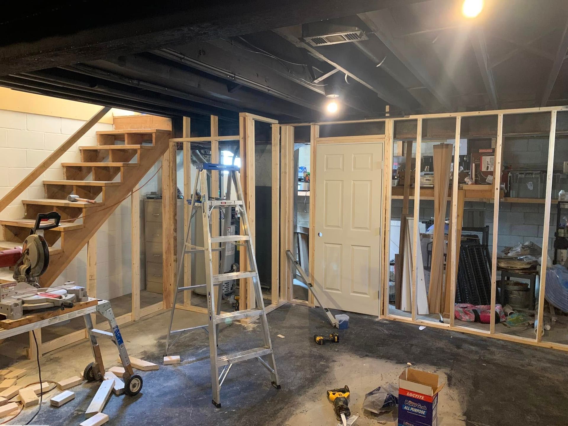 Framed wooden walls and an interior door under construction in a basement with exposed ceiling joists and a staircase.